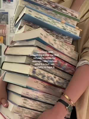 A person holds a large stack of books, likely textbooks, in their arms, with the books angled slightly towards the viewer. The person is wearing a light-colored blouse and a watch with a black strap and gold details. The background is blurred, appearing to be a room with partially visible wall decor. The overall color palette is muted with tones of beige and brown from the books and shirt.