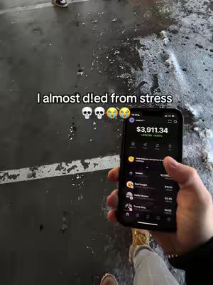 A person holds a smartphone displaying a financial app's interface. The background shows a wet, gritty surface and a person's legs and shoes. Text overlaid on the image reads: 'I almost died from stress', accompanied by emoji faces reflecting stress.