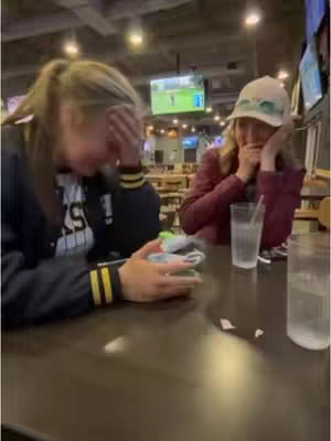 Two young women seated at a table in a bar setting, both looking upset with their hands covering their faces. The left person is wearing a dark jacket with yellow accents overlaying a white shirt with black text. The right person is wearing a hat and a dark red jacket. A glass of water and a straw are on the table.