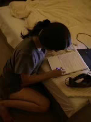 A person kneels on the floor in a softly lit room, bent over a notebook placed on a bed. The person has dark hair pulled back, concealing their face. A laptop and other items are visible, suggesting a study setup. The color palette is warm, dominated by the light tones of a bed and the shadow of the person.