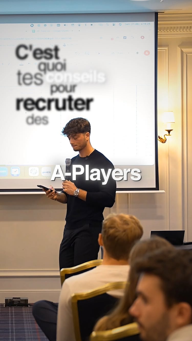 A man in a suit is standing on a stage in front of a screen, holding a phone and speaking to an audience. The screen shows a blurred text in French "C'est quoi tes conseils pour recruter des A-Players". The background appears to be a hotel or conference room.