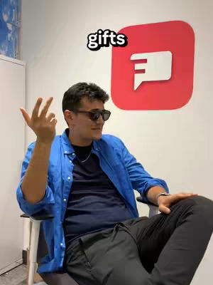 A man wearing sunglasses and a blue shirt sits in an office chair, making a hand gesture. Behind him is a large red logo with a white speech bubble icon. The word 'gifts' is overlaid at the top.