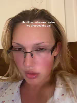 A selfie taken from the chest up of a fair-skinned woman with blonde hair, and black rimmed glasses. She appears to be lying down, with a light colored floral top on, and a light-colored backdrop. Text saying 'this filter makes me realise i've dropped the ball' appears on the image.