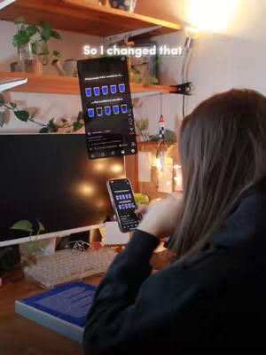 A person is sitting at a desk, the angle is from behind. On the desk, there is a monitor, a keyboard. Additionally, there is a phone and another screen, suggesting a focus on app use or content display. The overall impression is one of productivity and digital content creation. The lighting give a cozy vibe and looks like it has been captured inside a home.