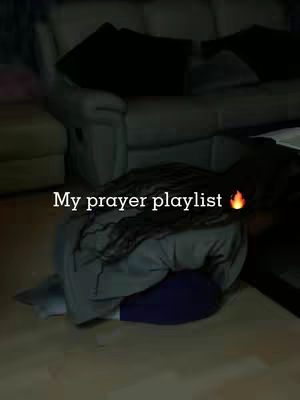 A person kneeling on the floor with their back to the camera, head bowed. The setting appears to be a living room with a sofa in the background. Text overlayed on the image says, 'My prayer playlist' with a fire emoji. The lighting is dim.