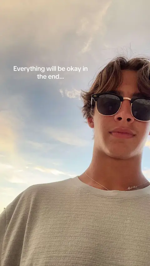 A close-up shot of a young man wearing sunglasses, looking towards the camera, with a sky background.