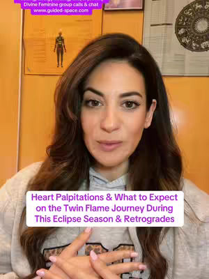 A woman with long brown hair faces the camera directly. She is wearing a light gray sweater over a gray shirt with a printed design. A white rectangular box with black text is positioned near her chin, and elements on the background include a panel with words and astrology symbols.