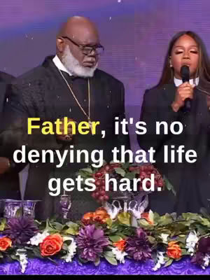 A medium shot shows a bald Black man in a black suit and glasses to the left, looking down. A fair-skinned Black woman stands to the right, holding a microphone and speaking. In the foreground, a table displays a floral arrangement with red, purple, and orange flowers.