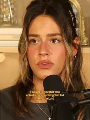 A woman with long, wavy brown hair and a serious expression is the focus. She is centered, with soft lighting and a neutral background. A black microphone is positioned in front of her, and text overlays are present at the bottom.