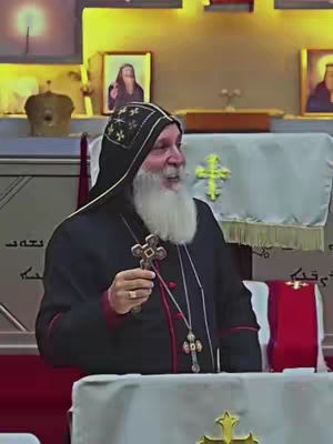 The image depicts a man in religious attire, likely a clergyman, holding a cross. He has a long white beard and is smiling. He is standing in what appears to be a church or religious setting, with religious art and decor visible in the background.