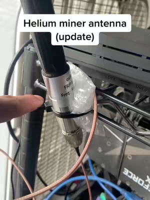 A close-up shot focuses on a metal antenna, connected with a series of cables and wires set against a cluttered background of metal shelving and electronic components. Text appears in a white box at the top, and a finger is touching the side of the antenna.