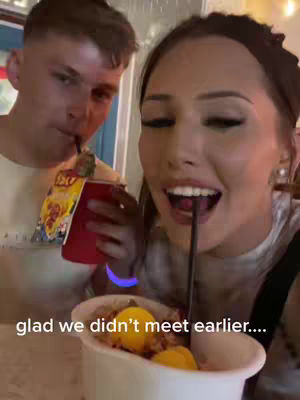 A selfie-style shot featuring a couple enjoying a meal together. The woman on the right is smiling and drinking from a straw, while the man on the left is also drinking from a straw, holding a red cup with yellow branding. A bowl of food is in front, containing yellow, round items, implying a dessert. The overall composition is casual and warm.