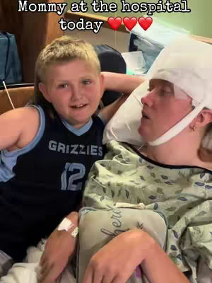 A young boy smiles while sitting next to a woman in a hospital bed. The woman is lying down, has a white bandage on her head, and is looking up with a concerned expression. The text overlay says 'Mommy at the hospital today' and includes three heart emojis.
