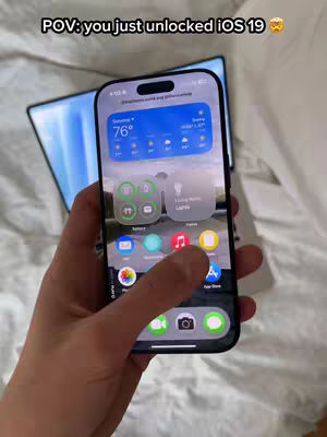 A hand holding and interacting with an iPhone. The iPhone screen displays a mock-up of iOS 19 with widgets and app icons. In the blurred background, a laptop is visible.