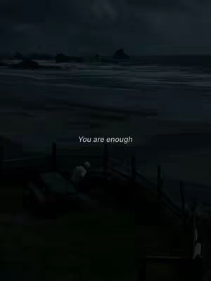 A dark, moody image of a beach at dusk. The sky is overcast, and the ocean is a deep shade of blue-gray. In the foreground, a fence and some figures are silhouetted against the fading light. Text overlayed on the image reads "You are enough".
