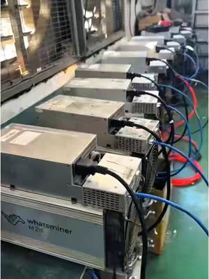 The image shows multiple Whatsminer M20S crypto mining machines lined up on a surface, likely in a warehouse or testing facility. They are silver in color with black cables and fans. The overall composition suggests a focus on the technology and the process of crypto mining hardware.