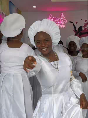 The image captures a group of women dressed in white robes and head coverings, smiling and pointing toward the camera, possibly in a religious setting. The composition is close, focusing on the expressions of the people.