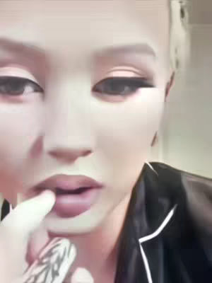 A close-up of a person's face, likely a woman, with pale skin, prominent dark eyeliner, and pink lips. She is touching her lower lip with her index finger, and a tattoo is visible on her left wrist. The background is plain and out of focus. The overall tone seems introspective.