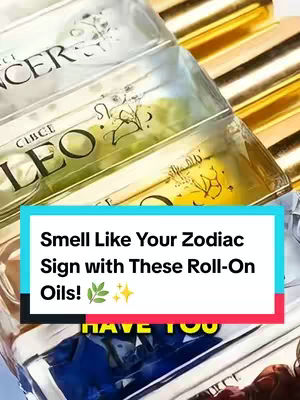 The image shows a collection of roll-on essential oil bottles, arranged in a slightly downwards diagonal on a surface. The bottles are clear with gold or silver lids, and each has a label with the name of a zodiac sign in a stylized font. The names 'Leo' and 'Cancer' are clearly visible. The background is out of focus, with a gradient of gold and yellow colors. A graphic element in the form of a blue rectangular box is placed at the bottom, containing text that reads 'Smell Like Your Zodiac Sign With These Roll-On Oils!' followed by two small green leaves and sparkles.