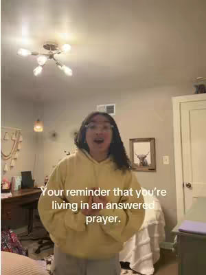 A woman with long dark hair and glasses stands in a room with her arms outstretched, looking at the camera. Text overlayed reads, 'Your reminder that you're living in an answered prayer.' The room has a bed, a desk, and a framed picture of a cow.