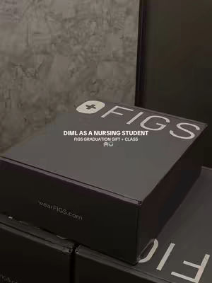 The image shows a dark gray FIGS box on top of another. The front of the top box has the white text reading 'FIGS' and 'DIML AS A NURSING STUDENT FIGS GRADUATION GIFT + CLASS'. The background is a textured gray wall.