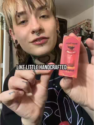 A woman with blonde hair and multiple piercings looks directly at the camera, holding a small, rectangular box with text. She is wearing a black top and has tattoos on her neck and hand. The background is a blurred interior space.