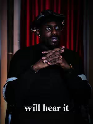 A man with dark skin and a goatee is the central focus, his hands clasped in front of him. He wears a black top and a black cap. The background consists of red draped fabric with vertical lines.