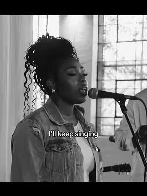 A black and white image of a woman singing into a microphone. She is wearing a denim jacket over a white top. The window is in the background. The text overlay says: I'll keep singing