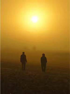Two silhouetted figures stand on a field, facing away from the camera, under a bright, glowing sun in a hazy atmosphere. The scene is dominated by warm, golden hues.