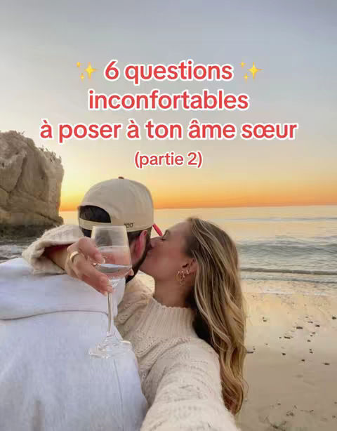 A selfie-style shot of a couple on a beach at sunset. A woman kisses the man's cheek while holding a glass of wine.