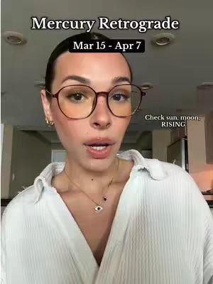 A woman with glasses is centered in a selfie-style shot. The background is a softly lit room. She is wearing a white collared shirt and a gold necklace with a charm. The image is well-lit, creating a bright and engaging visual.