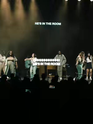 The cover image shows a live performance on a stage, dimly lit with spotlights and a dark background. Several people are present on the stage, with some in the foreground and more in the background. In the foreground, a group of people are standing close to each other. Above them, on the stage curtain, the words "HE'S IN THE ROOM" are written in bright green letters. Below, there's an additional text overlay consisting of the words "OHHHHHHHHHHHH HE'S IN THE ROOM". The overall atmosphere suggests a worship or performance setting.