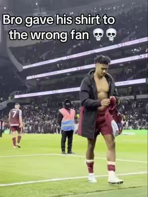 A man stands shirtless in a stadium, holding a shirt. Text overlay suggests humor about the shirt. The background shows a crowd. Colors include green grass and white seating.