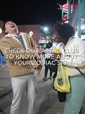 An eye-level outdoor shot captures two individuals engaged in a street interview. The focus is on two people, one is a man in a tan jacket who is laughing, and the other is a woman with glasses and long hair, holding a microphone, and a yellow shopping bag.