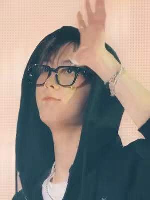 A person with dark hair, wearing black-framed glasses and a black hoodie, is reaching up with their right hand. The background is a blurred peachy-pink.