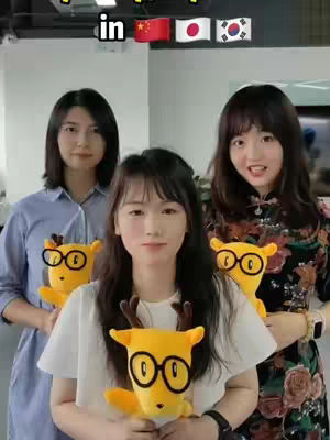 Three young Asian women are featured in a well-lit office setting. They stand side-by-side, holding yellow stuffed animals adorned with glasses. The backdrop is a plain, light-colored wall. The women are dressed in casual, everyday attire. Overlayed above them is text that says "in" with flags of China, Japan and Korea.