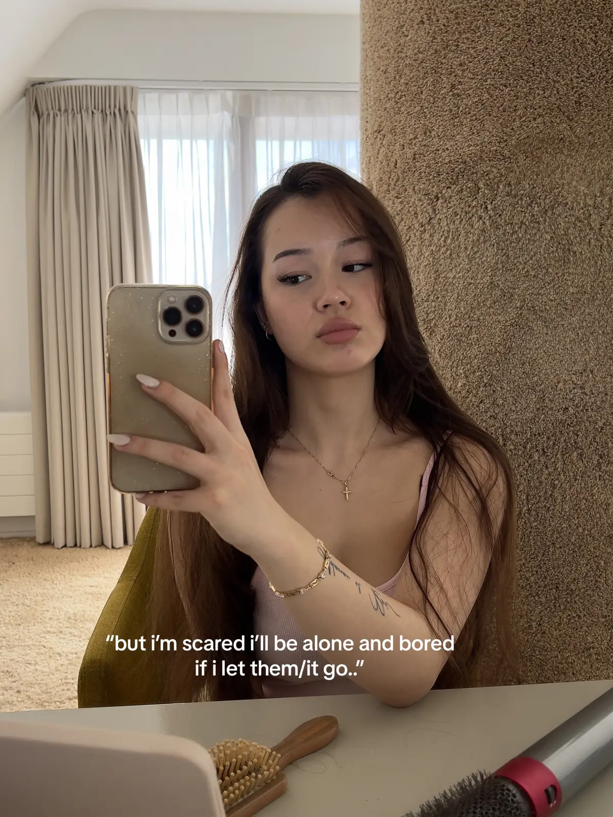 A young woman taking a selfie with a phone, she is looking down and to her left, the background consists of curtains, a textured wall, and a desk.