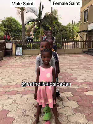 A photo taken from the ground level presents a group of school-aged children lined up in a row. There are two distinct vertical text labels, each positioned above the children's heads: one reads 'Male Saint' and the other reads 'Female Saint', delineating the gender division in the line. The children are well-lit, with the central focus on a girl in the center, dressed in a pink dress with white polka dots, playfully positioned above a green toy. The background consists of vibrant brick patterns, shrubbery, and a building, all contributing to a bright and dynamic setting.