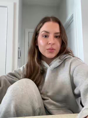 A woman is seated, facing forward, with neutral expression, wearing a grey hoodie. The background shows a lit room, with white walls and a closed door.