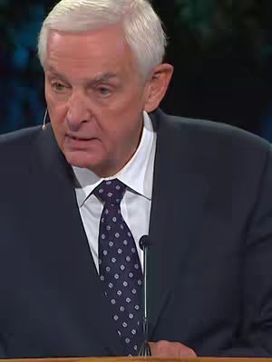 A medium shot of an older man wearing a dark suit, white collared shirt, and a patterned tie. He is speaking into a microphone, with a focused expression. The background is slightly blurred, possibly a stage or studio.