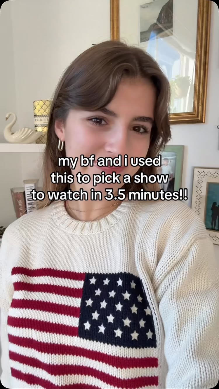 A young woman with light brown hair, wearing a white sweater with a large American flag design, is the central focus of the image. Text overlays describe how she used something to show a show to watch in 3.5 minutes. The background includes parts of a room, like a wall and some framed pictures. The woman is smiling at the camera.