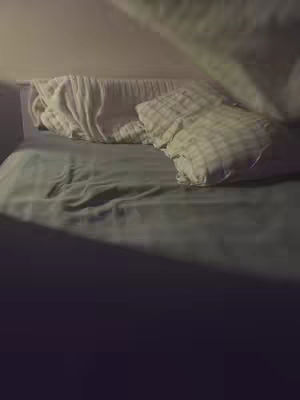 The image shows a dimly lit bedroom scene with a bed taking up most of the frame. The bed has a white headboard and is covered with a gray sheet and pillows. A blanket is being lifted or draped over the bed, adding a feeling of intimacy. The lighting suggests a time of day or setting and that everything is very quiet and peaceful.