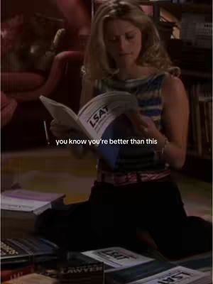 A woman sits on the floor surrounded by books, reading a large book with 'LSAT' printed on one of its pages. She's wearing a striped top and black pants, and the background includes a dimly lit room with a red armchair and additional books.