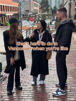 An outdoor shot featuring three people. A person on the left is holding a microphone and facing two other people. The person in the middle is holding the microphone and speaking, while the person on the right is listening. The background shows a city street with buildings and a stop sign.