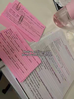 The image displays a top-down view of a desk with pink and white papers covered in handwritten notes, a pink water bottle, and a pen. Text overlayed on the image reads, 'Only a genius could love a woman like me.' The composition suggests a study or workspace, with the papers and bottle possibly indicating study materials and a drink.