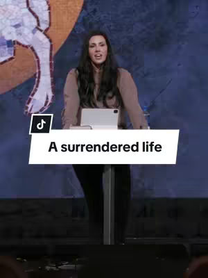 A woman stands behind a lectern with a tablet, speaking into a microphone. Behind her is a blurred, abstract image in shades of blue and gold. A white banner with black text stating 'A surrendered life' overlays part of the image.