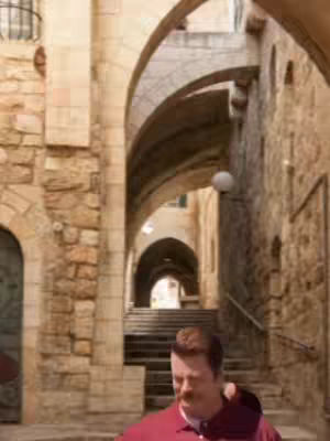 The image features a man with a mustache standing in a stone alleyway. The composition is framed by archways and staircases leading up, with the overall color palette being dominated by earthy tones.