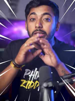 A man with dark hair and a beard is centered in the frame, looking directly at the viewer. He is wearing a black t-shirt with white and yellow lettering. His hands are clasped together near his face, and a microphone is positioned in front of him. Behind him, the background suggests a studio setting with lighting and a purple screen.