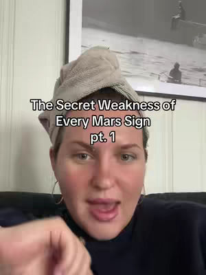 A woman with fair skin is centered in the frame, wearing a dark top and a head wrap. The text overlay reads, The Secret Weakness of Every Mars Sign pt. 1, the image is lit with natural light, and a black and white framed picture hangs in the background.