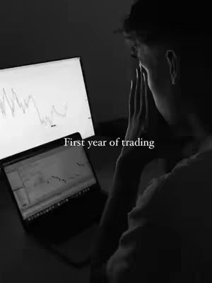 A grayscale image features a person seated, facing away from the viewer, with their head resting on their hands. Two computer screens display financial charts, and a laptop is open. The scene is dimly lit, creating a somber mood. Text overlayed on the image reads 'First year of trading.'